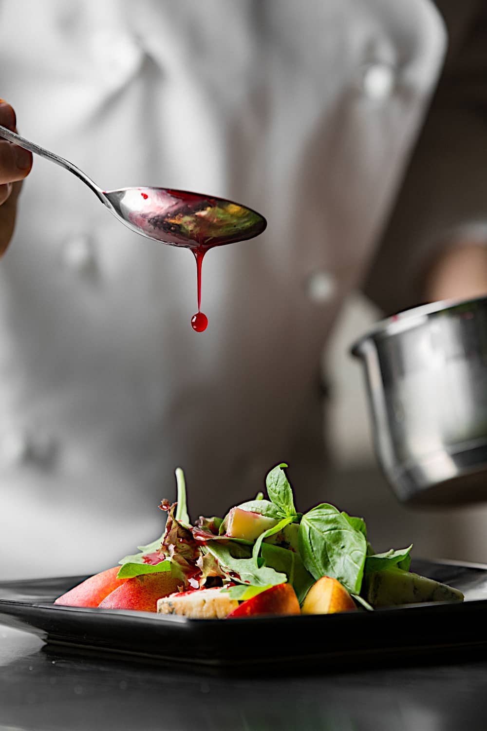 Chef drizzles red dressing onto a colorful salad on a black plate, greens and fruit visible, in a professional kitchen.
